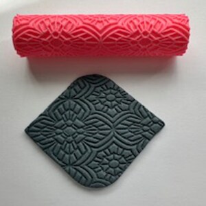 May include: A red patterned rolling pin and a grey textured square. The rolling pin has a repeating floral design. The grey square has a similar floral pattern. The items are likely for crafts or baking.