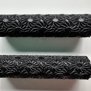 May include: Two black textured rollers with a repeating floral pattern. The rollers are cylindrical and appear to be made of a rubber or similar material. The floral design is raised, creating a tactile surface.