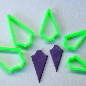 Puede incluir: Un juego de seis cortadores de galletas de plástico verde en forma de puntas de flecha, y dos puntas de flecha de fondant morado.