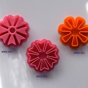Puede incluir: Tres cortadores de galletas con forma de flores. El cortador superior es rosa y está etiquetado como MMS-001. El cortador inferior izquierdo es rosa y está etiquetado como MMS-002. El cortador inferior derecho es naranja y está etiquetado como MMS-003.