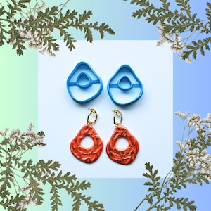 Puede incluir: Un par de pendientes abstractos naranjas con aros dorados, junto a dos cortadores de pendientes azules. Los pendientes tienen una superficie texturizada y un agujero en el centro. El fondo es blanco y degradado azul y verde, con detalles florales.