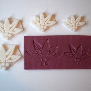 Peut inclure: Cinq tampons en silicone blanc avec un motif floral, comprenant un motif de feuilles et de baies. Les tampons sont disposés sur une surface blanche à côté d'une feuille de silicone bordeaux avec deux des mêmes motifs floraux.