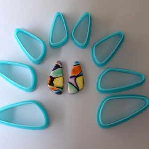 Op de afbeelding: Een set van acht turquoise blauwe plastic mallen in de vorm van een traan, gerangschikt in een cirkel rond twee kleurrijke, abstracte, traanvormige polymer clay oorbellen.