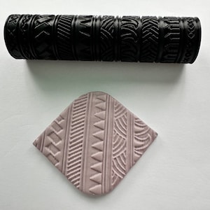 Puede incluir: Un rodillo negro con una variedad de patrones geométricos tallados en la superficie. El rodillo se utiliza para crear patrones decorativos en arcilla o masa. Una muestra del patrón se muestra en la imagen, que es de color morado claro.