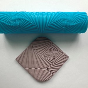 Könnte beinhalten: Eine blaue Teigrolle mit einem geometrischen Muster und einer Probe des Musters, das in grauen Ton geprägt ist.
