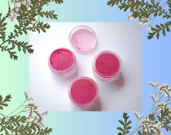 Puede incluir: Cuatro pequeños frascos transparentes de maquillaje. Tres frascos contienen un polvo rosa vibrante, mientras que el cuarto contiene un tono rosa más claro. Los frascos están dispuestos sobre una superficie blanca, con un fondo degradado azul y verde. Acentos florales enmarcan la imagen.