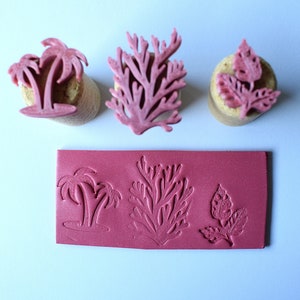 May include: A collection of pink rubber stamps featuring botanical designs. The stamps include palm trees, coral, and leaf patterns. The image also shows the stamped impressions of these designs on a pink rectangular surface.
