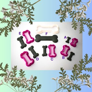 May include: A selection of dog bone-shaped cookie cutters and biscuits. The cutters are pink and white, while the biscuits are dark grey. The arrangement includes various sizes and styles, ideal for baking treats.