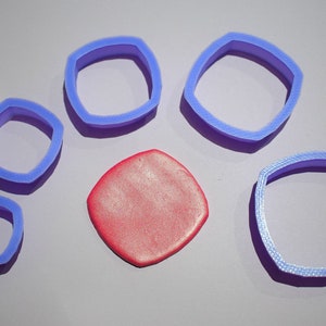 Op de afbeelding: Vijf verschillende maten blauwe plastic vierkante koekjesvormen met afgeronde hoeken. In het midden van de afbeelding bevindt zich een vierkant stuk modelleerklei in rood.
