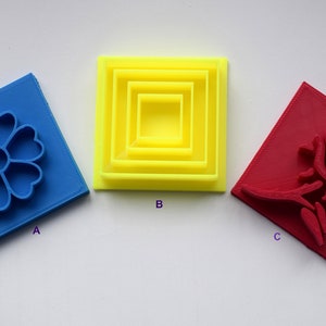 May include: Three 3D printed cookie cutters, one blue with a flower design, one yellow with a set of nested squares, and one red with a rose design.