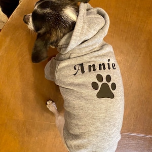 K&ouml;nnte beinhalten: Ein grauer Kapuzenpullover f&uuml;r einen Hund mit dem Namen "Annie" und einem schwarzen Pfotenabdruck auf der R&uuml;ckseite.