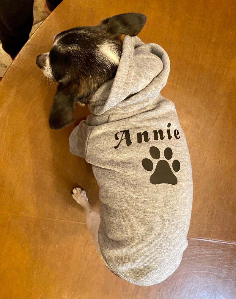 K&ouml;nnte beinhalten: Ein grauer Kapuzenpullover f&uuml;r einen Hund mit dem Namen "Annie" und einem schwarzen Pfotenabdruck auf der R&uuml;ckseite.