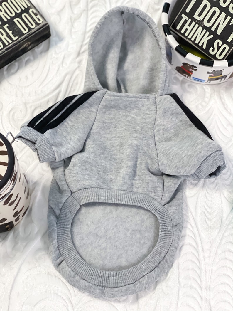 K&ouml;nnte beinhalten: Ein grauer Kapuzenpullover mit schwarzen Streifen an den &Auml;rmeln. Der Pullover ist f&uuml;r einen Hund konzipiert und hat ein Loch f&uuml;r den Schwanz.