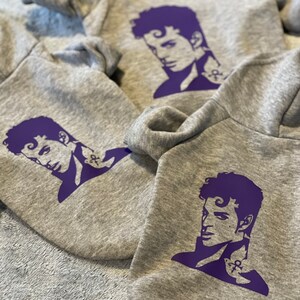K&ouml;nnte beinhalten: Drei graue Kapuzenpullover mit einer lilafarbenen Silhouette von Prince auf der Vorderseite. Jeder Pullover zeigt eine andere Pose von Prince.