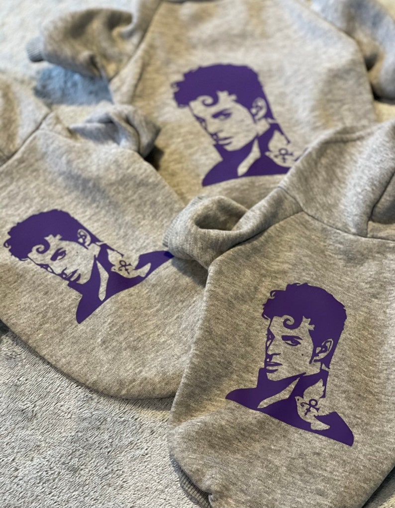 K&ouml;nnte beinhalten: Drei graue Kapuzenpullover mit einer lilafarbenen Silhouette von Prince auf der Vorderseite. Jeder Pullover zeigt eine andere Pose von Prince.