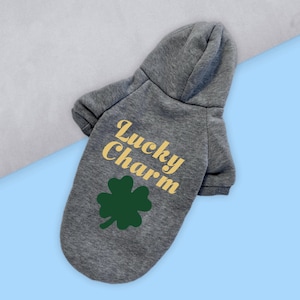 Op de afbeelding: Een grijze hoodie voor honden met de tekst "Lucky Charm" in geel en een groene klaver met vier bladeren.
