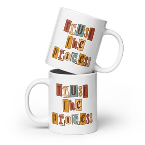 May include: Two white ceramic mugs stacked on top of each other. Both mugs feature the phrase "TRUST the PROCESS" in colorful, retro-style letters.