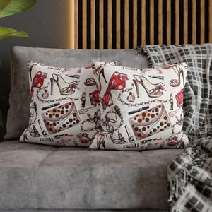 Op de afbeelding: Twee decoratieve kussens met een witte achtergrond en een herhalend patroon van hoge hakken, handtassen, lippenstift en parfumflesjes in rood, bruin en zwart. De kussens liggen op een grijze bank met een patroon deken.
