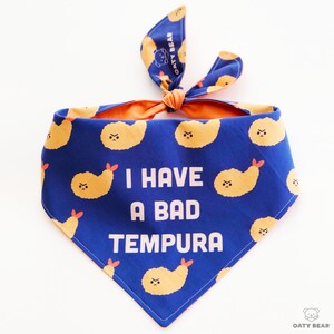 May include: Blue bandana with a white print that says "I have a bad tempura". The bandana is decorated with a pattern of yellow cartoon tempura shrimp with angry faces.
