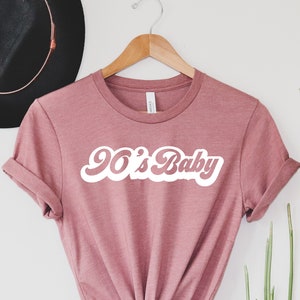 May include: A mauve-colored t-shirt with the text "90's Baby" in a white, retro font. The shirt is hanging on a wooden hanger against a white background. A black hat is visible in the background.