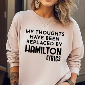 May include: A light pink long-sleeved sweatshirt with the text "My thoughts have been replaced by Hamilton lyrics" printed in black.