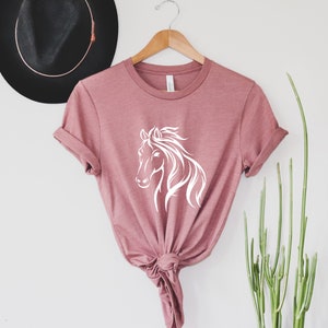 Peut inclure: Un t-shirt rose poussi&eacute;reux avec un graphique de silhouette de cheval blanc. Le t-shirt est nou&eacute; &agrave; l'avant et a les manches retrouss&eacute;es.