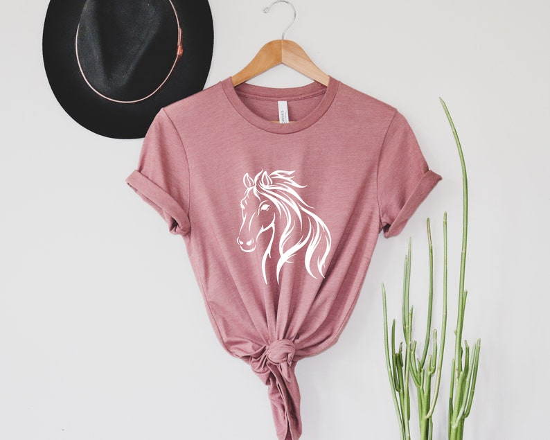 Peut inclure: Un t-shirt rose poussi&eacute;reux avec un graphique de silhouette de cheval blanc. Le t-shirt est nou&eacute; &agrave; l'avant et a les manches retrouss&eacute;es.