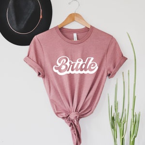 K&ouml;nnte beinhalten: Ein mauvefarbenes T-Shirt mit dem Wort "Bride" in wei&szlig;en Retro-Lettern.