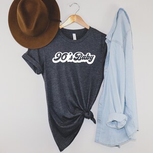 May include: Dark gray t-shirt with the text "90's Baby" in white script. A brown hat hangs to the left, and a light blue denim shirt is draped on the right. The t-shirt is knotted at the bottom.