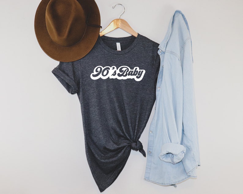 May include: Dark gray t-shirt with the text "90's Baby" in white script. A brown hat hangs to the left, and a light blue denim shirt is draped on the right. The t-shirt is knotted at the bottom.