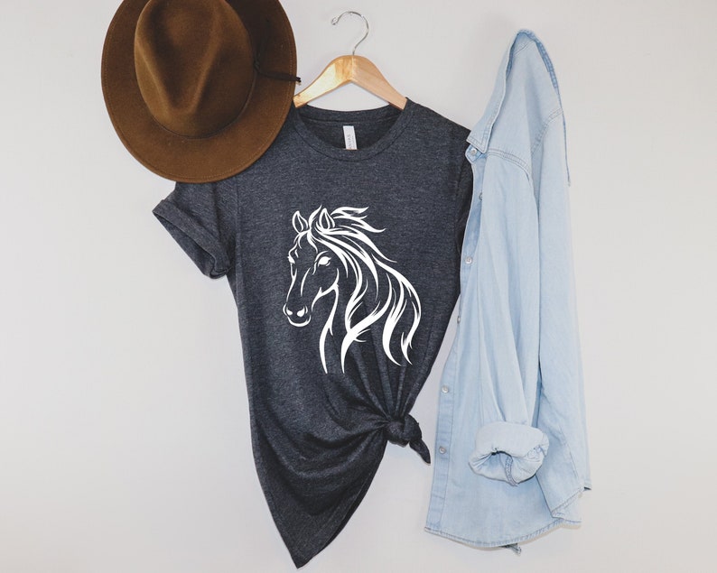 Peut inclure: Un t-shirt gris fonc&eacute; avec une silhouette blanche de t&ecirc;te de cheval. Le t-shirt est pr&eacute;sent&eacute; sur un fond blanc avec un chapeau marron et une chemise en jean bleu clair.