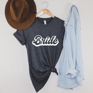 K&ouml;nnte beinhalten: Ein dunkelgraues T-Shirt mit dem Wort "Bride" in wei&szlig;er Retro-Schrift. Das Shirt ist in der Taille gebunden und wird mit einem hellblauen Jeanshemd kombiniert.