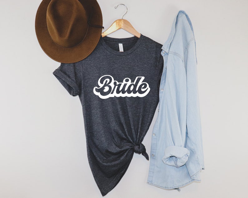 K&ouml;nnte beinhalten: Ein dunkelgraues T-Shirt mit dem Wort "Bride" in wei&szlig;er Retro-Schrift. Das Shirt ist in der Taille gebunden und wird mit einem hellblauen Jeanshemd kombiniert.