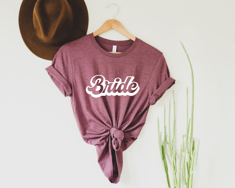 K&ouml;nnte beinhalten: Ein maroonfarbenes T-Shirt mit dem Wort "Bride" in wei&szlig;er Retro-Schrift.