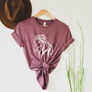 Peut inclure: Un t-shirt marron avec un motif de silhouette de cheval blanc. Le t-shirt est nou&eacute; en bas et suspendu &agrave; un cintre en bois.