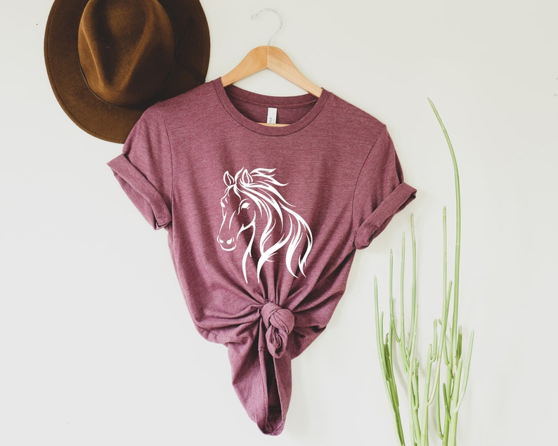 Peut inclure: Un t-shirt marron avec un motif de silhouette de cheval blanc. Le t-shirt est nou&eacute; en bas et suspendu &agrave; un cintre en bois.