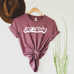 May include: Maroon t-shirt with the text "90's Baby" in a white, retro font. The shirt is knotted at the waist and hangs on a wooden hanger. A brown hat is visible in the background, along with green plant stems.