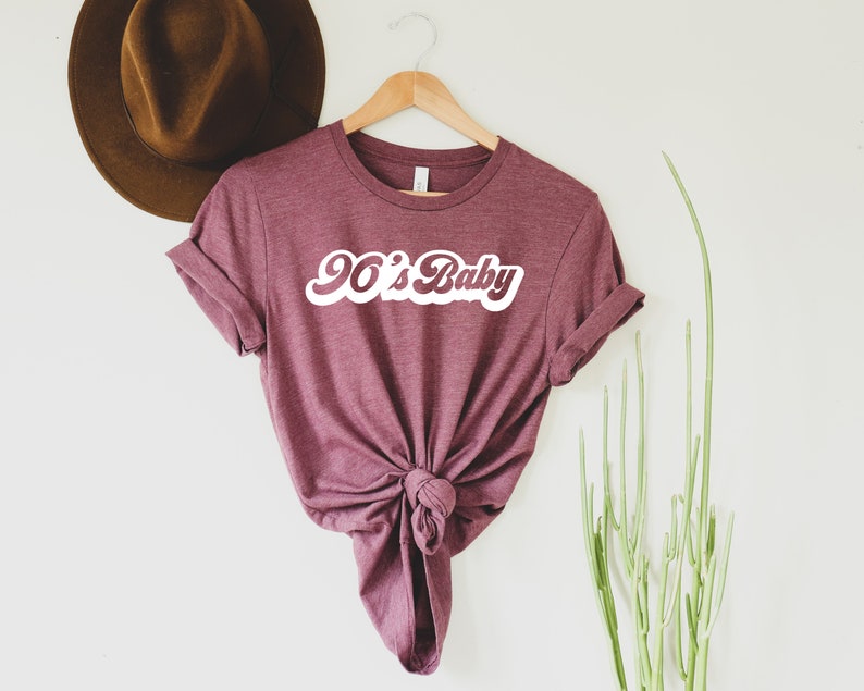 May include: Maroon t-shirt with the text "90's Baby" in a white, retro font. The shirt is knotted at the waist and hangs on a wooden hanger. A brown hat is visible in the background, along with green plant stems.
