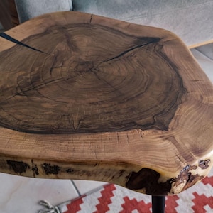 May include: A live edge side table featuring a circular wooden slab top with a dark brown center and lighter brown outer rings. The wood grain is visible, showing natural cracks and knots. The table is supported by thin black metal legs.