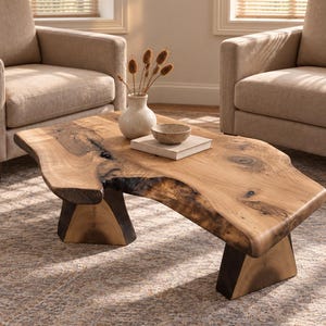 May include: A live-edge wooden coffee table with a natural grain pattern and dark knots, supported by two thick, angular wooden legs. On the table sits a light-colored ceramic vase with dried stems, a small speckled bowl, and a rectangular book.