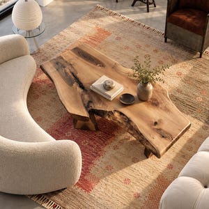 May include: A live-edge wooden coffee table with a natural grain and dark knots, featuring a smooth surface. On the table sits a stack of two white books, a small round stone object, a dark bowl, and a ceramic vase with green leafy branches.