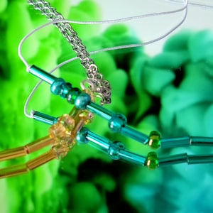 May include: Two necklaces with different coloured beads. One necklace has gold beads and the other has teal beads. Both necklaces are on a white surface with a green and blue background.