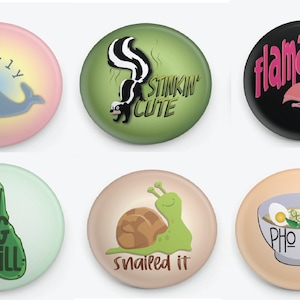 May include: Six round button pins with various illustrations and text. The pins feature a narwhal, a skunk, a flamingo, a pickle, a snail, and a bowl of pho. The text on the pins reads "narly", "stinkin' cute", "flamazing", "pretty big dill", "snailed it", and "pho real".