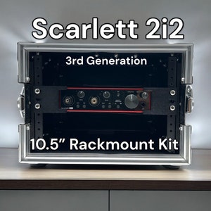 Peut inclure: Un kit de montage en rack argenté de 10,5 pouces avec une interface audio noire à l'intérieur. L'interface audio est étiquetée "Scarlett 2i2 3ème génération".