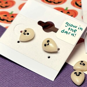 May include: Off-white ghost-shaped earrings with black eyes and smiles, displayed on white cards. The cards have a brown earring holder and a small tag that says "glow in the dark!". The background features a purple surface and a Halloween-themed pumpkin pattern.