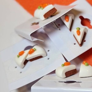 May include: A set of four pairs of carrot cake slice earrings. The earrings are made of polymer clay and are painted to look like carrot cake. Each earring features a slice of cake with a white frosting and a small orange carrot on top. The earrings are displayed on white card stock.
