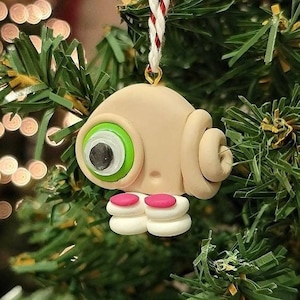 May include: A handmade Christmas ornament shaped like a cartoon character with a green eye, pink shoes, and a beige body. The ornament is hanging on a green artificial Christmas tree.