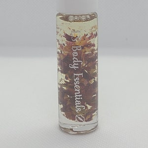 Op de afbeelding: Een transparante glazen rollerfles met een witte dop, gevuld met een lichtgele vloeistof en gedroogde bloemblaadjes. De fles heeft een etiket met de tekst "Body Essentials".