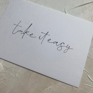 Puede incluir: Tarjeta blanca con la frase manuscrita "take it easy" en tinta negra.