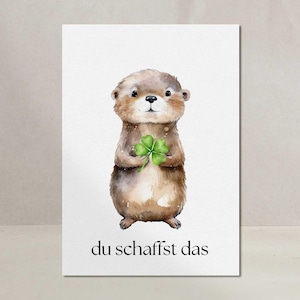 Könnte beinhalten: Aquarellillustration eines braunen Otters, der ein grünes vierblättriges Kleeblatt hält. Der Otter hat einen süßen Ausdruck und steht auf weißem Hintergrund. Der Text "du schaffst das" ist unten in Schwarz gedruckt.